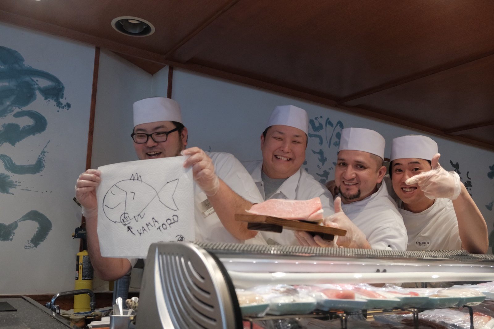 Four sushi chefs grinning at the counter, one holding a hand-drawn fish
