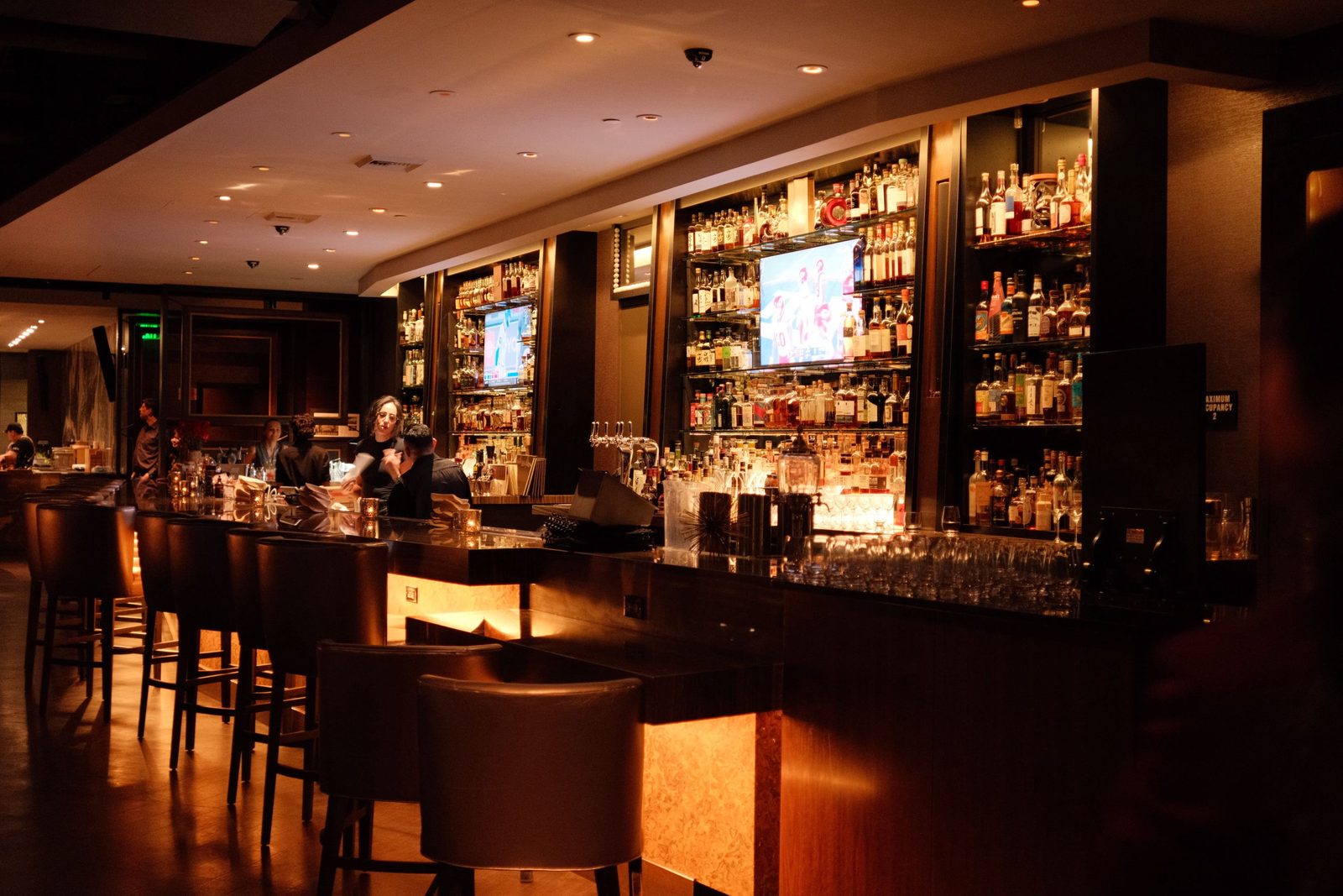 A warm, dim bar with backlit liquor shelves and leather stools