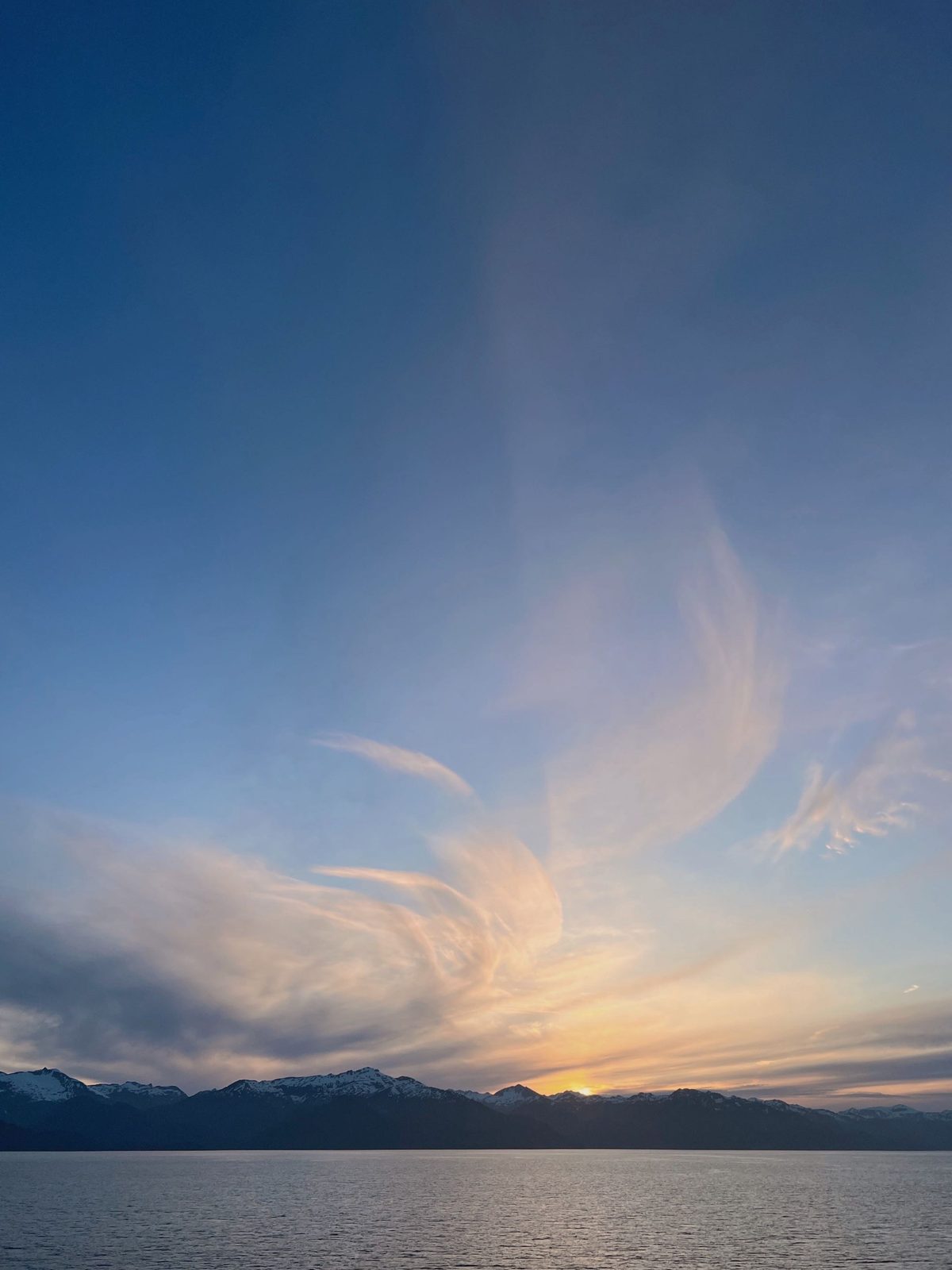 Wispy clouds over snow-capped peaks at sunset, seen from the water