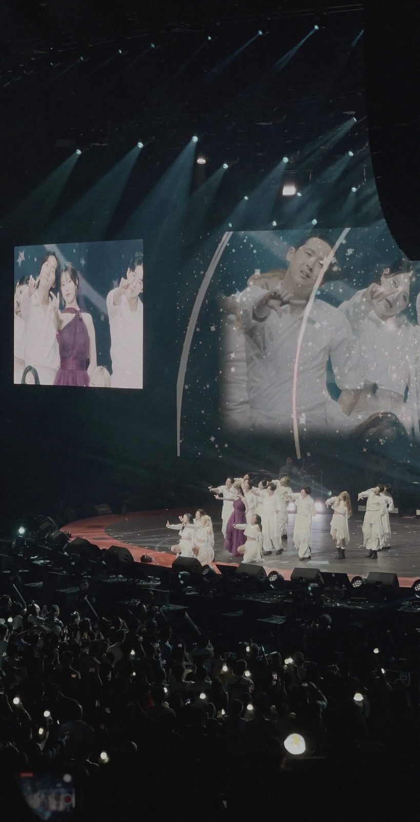 IU in a deep purple gown performing at the HEREH World Tour, surrounded by dancers in white on a curved stage with close-ups on the screens above