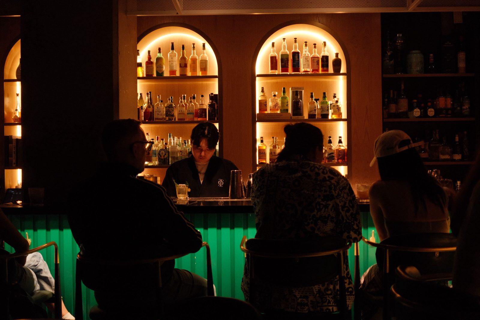 A bartender at work in a dim bar with backlit arched shelves