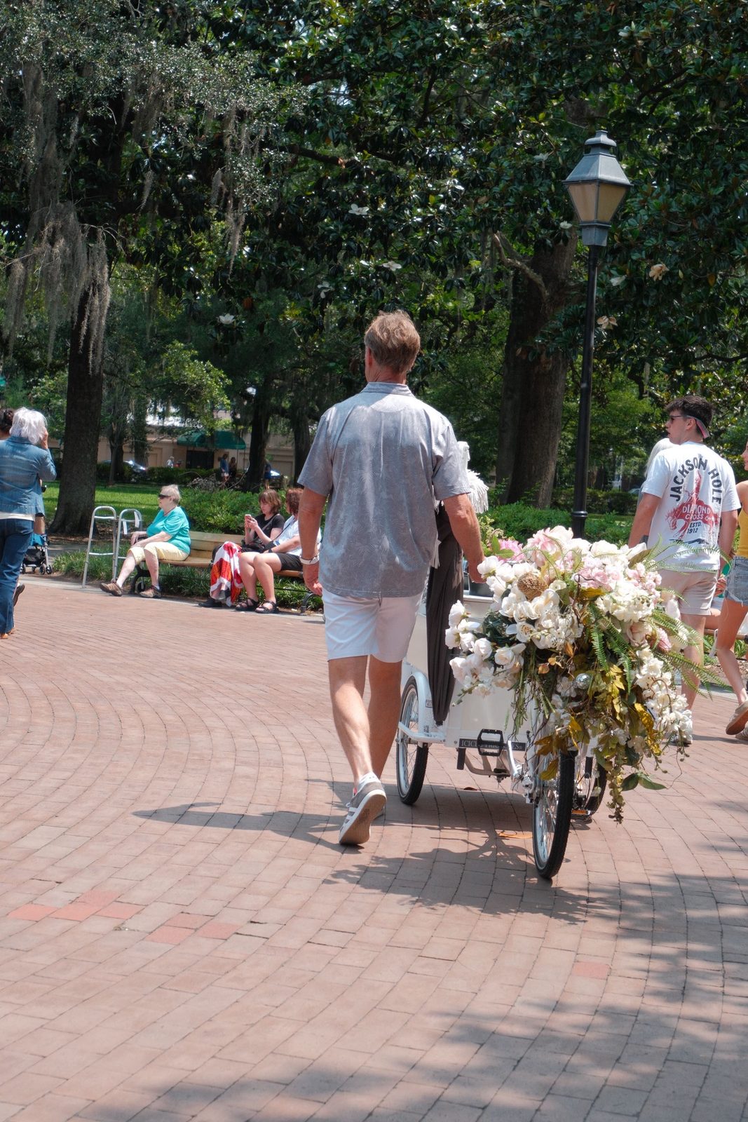 A man pushing a flower-laden cart through a tree-lined park