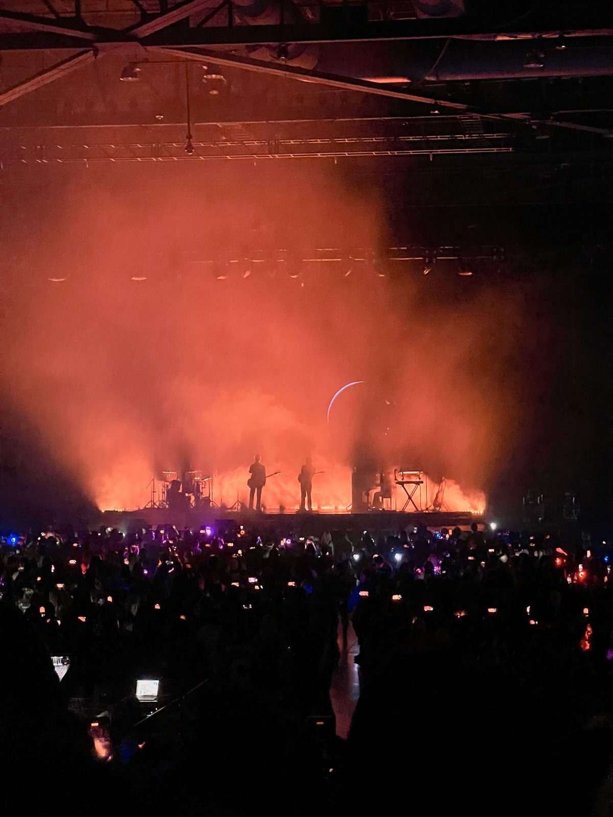 A band silhouetted against red smoke and a crescent moon, phone lights in the crowd