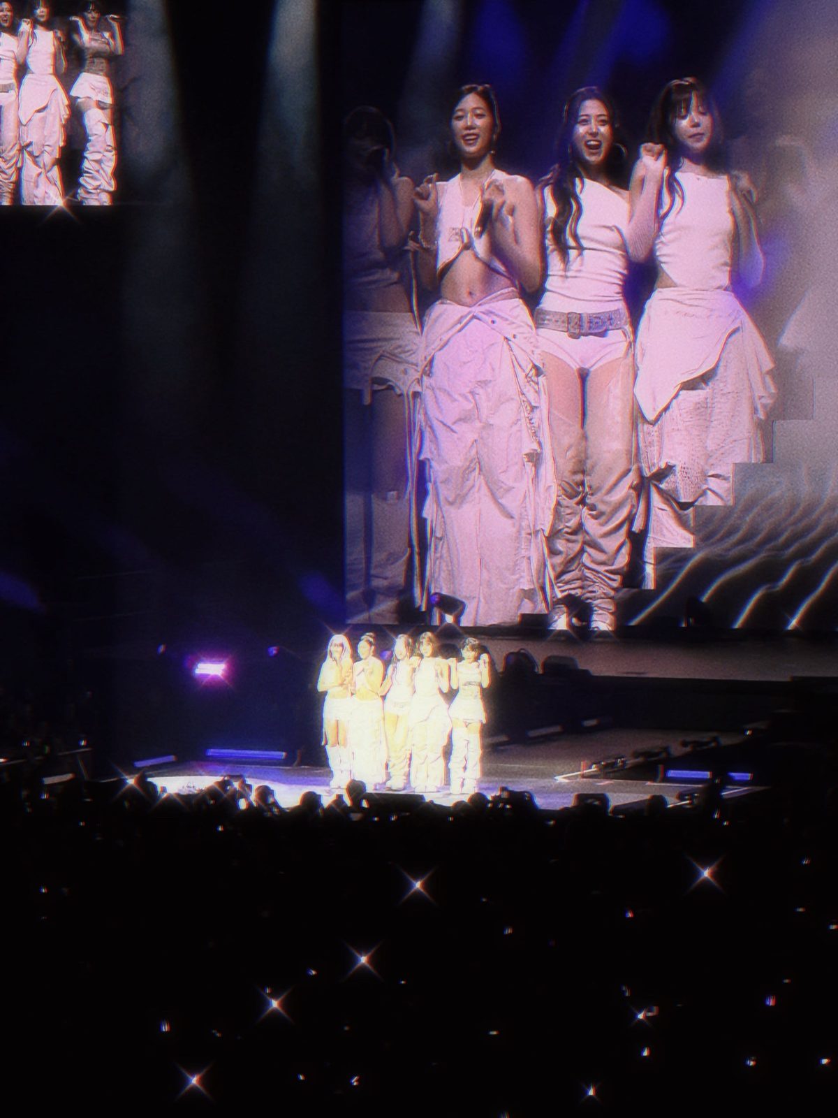 A girl group in white on stage, smiling close-up on the big screen above