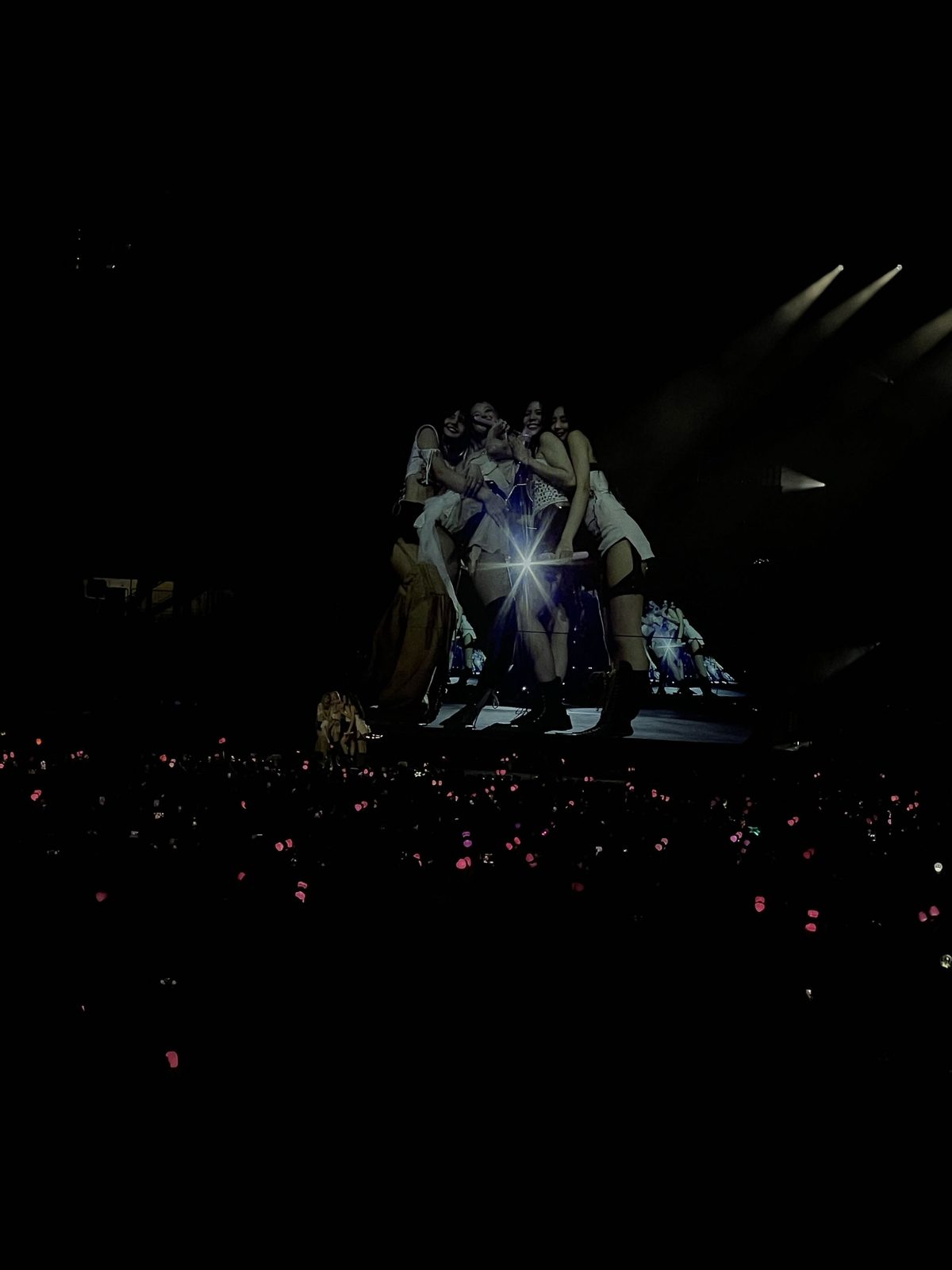 Performers huddled together on a dark stage above a sea of pink lights