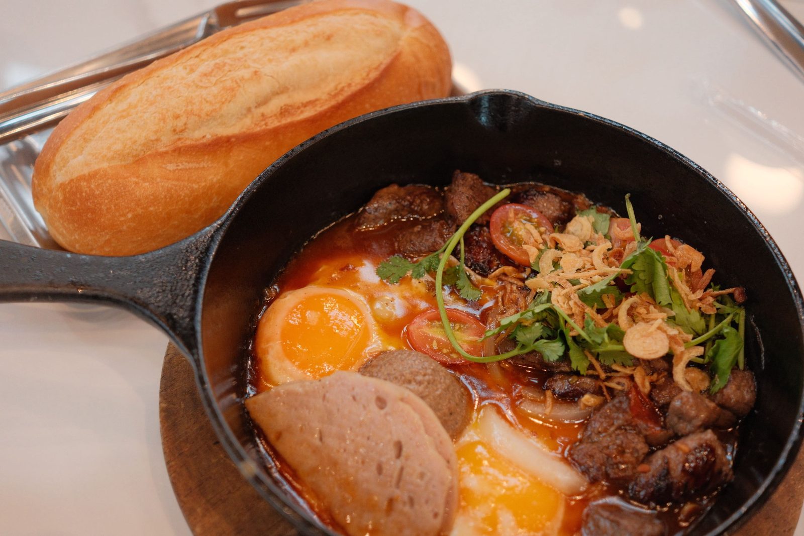 A sizzling skillet of beef stew and eggs served with a crusty bread roll